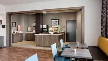 A modern hotel room with a kitchenette, a dining area with tables and chairs, and a separate entrance door.