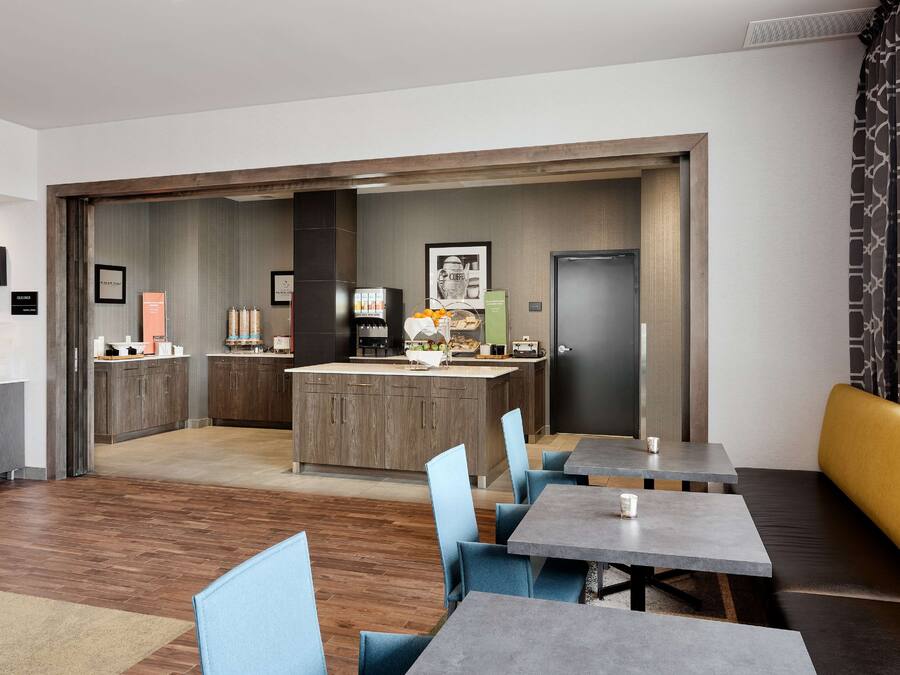 A modern hotel room with a kitchenette, a dining area with tables and chairs, and a separate entrance door.
