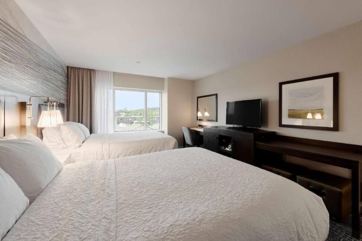 A hotel room with two beds, a desk, a TV, and a window with a view.