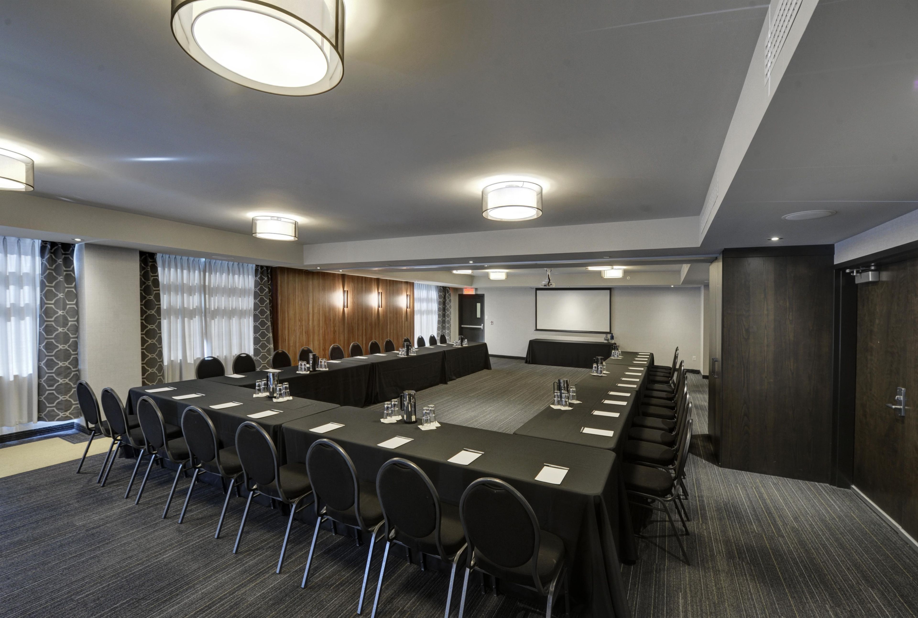 Un salón de conferencias con una larga mesa, sillas, una pantalla de proyector y una pantalla para presentaciones.