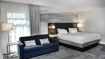 Quarto de hotel com uma cama grande, um sofá azul escuro, uma luminária moderna e vista através de cortinas translúcidas.