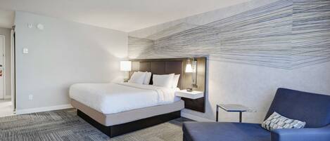 A modern hotel room with a large bed, a desk with a lamp, a blue armchair, and a patterned wall.