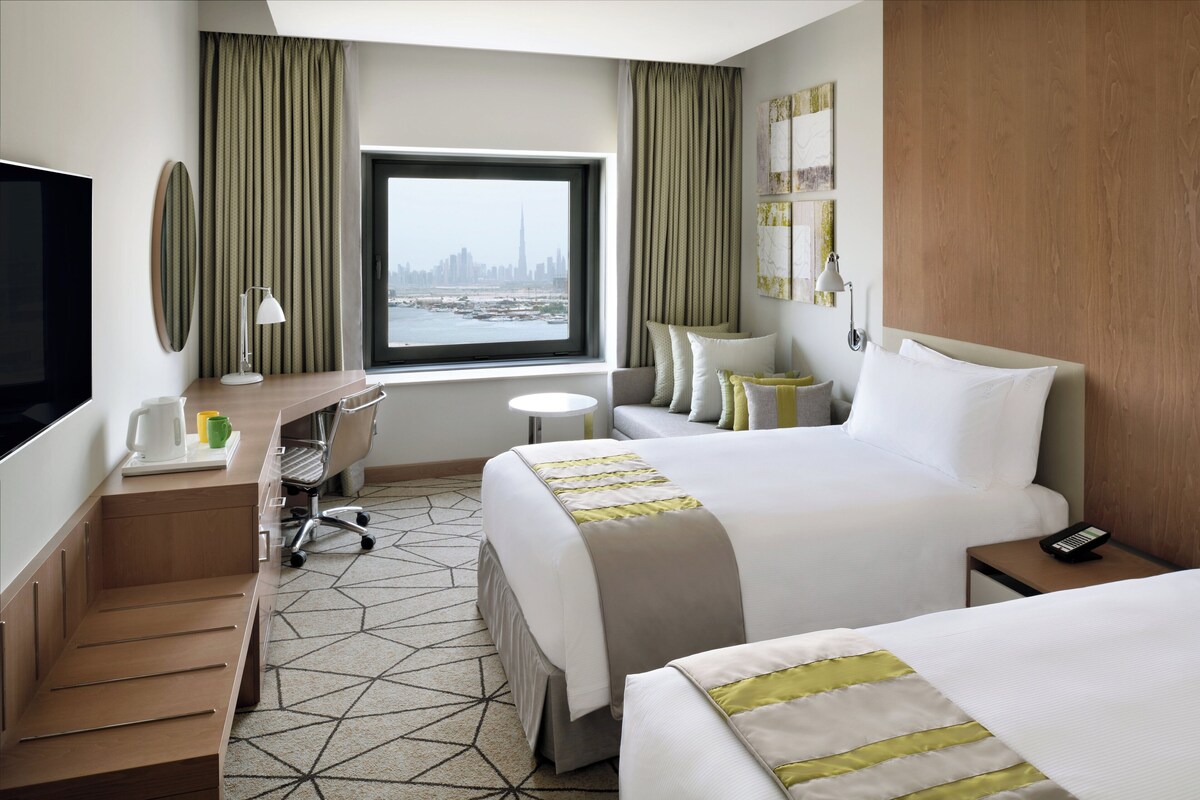 Une chambre d’hôtel équipée d’un lit, d’un bureau, d’une chaise, d’une télévision, d’une fenêtre donnant sur la ville et d’un téléphone sur la table de chevet.