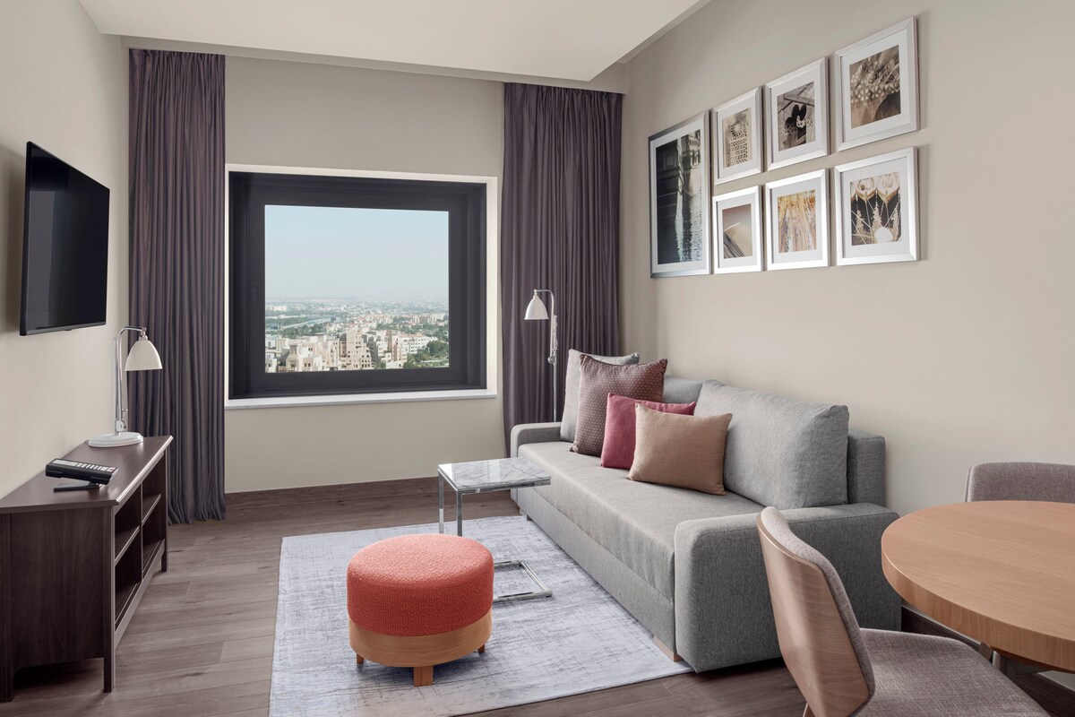 Un salon moderne avec un canapé gris, une table ronde en bois, un pouf rouge, un téléviseur et des tableaux encadrés accrochés au mur.