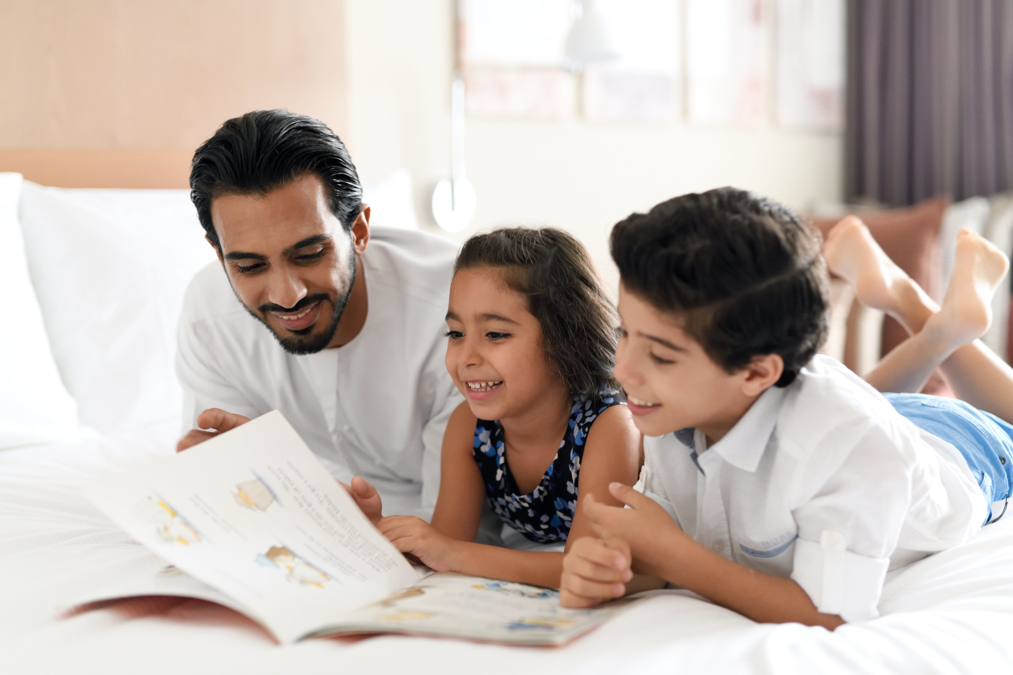 A family of three lying on a bed, reading a book together.