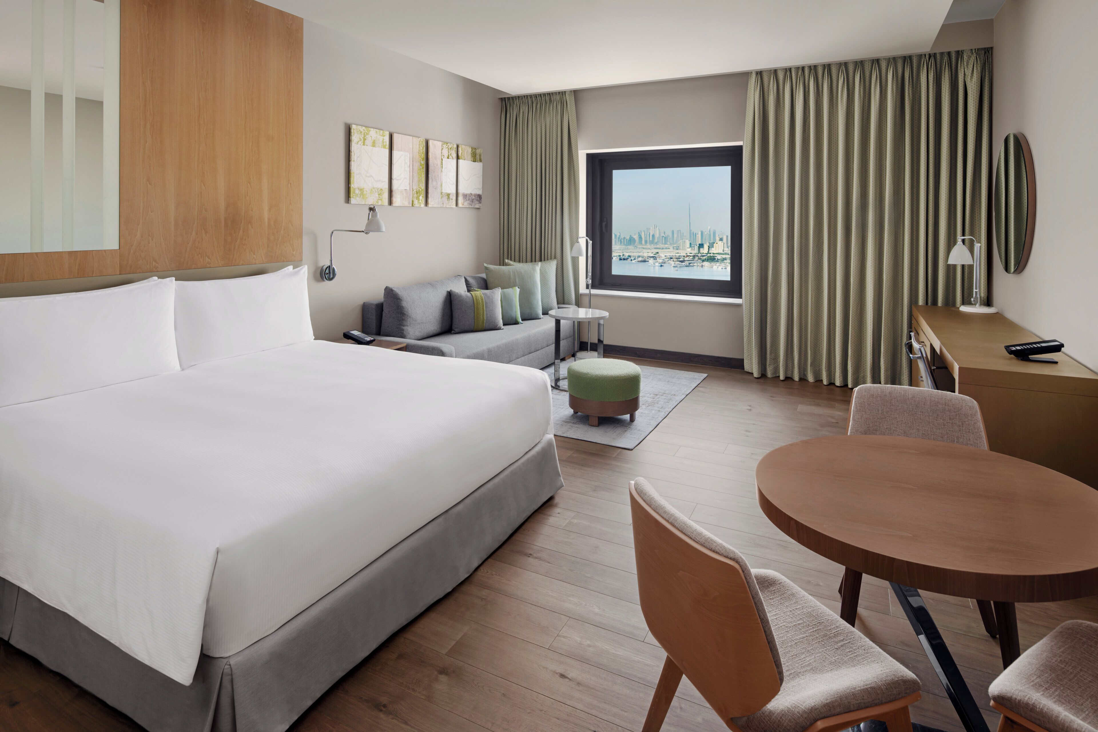 A hotel room with a large bed, a sofa, a dining table, and a view of a cityscape.