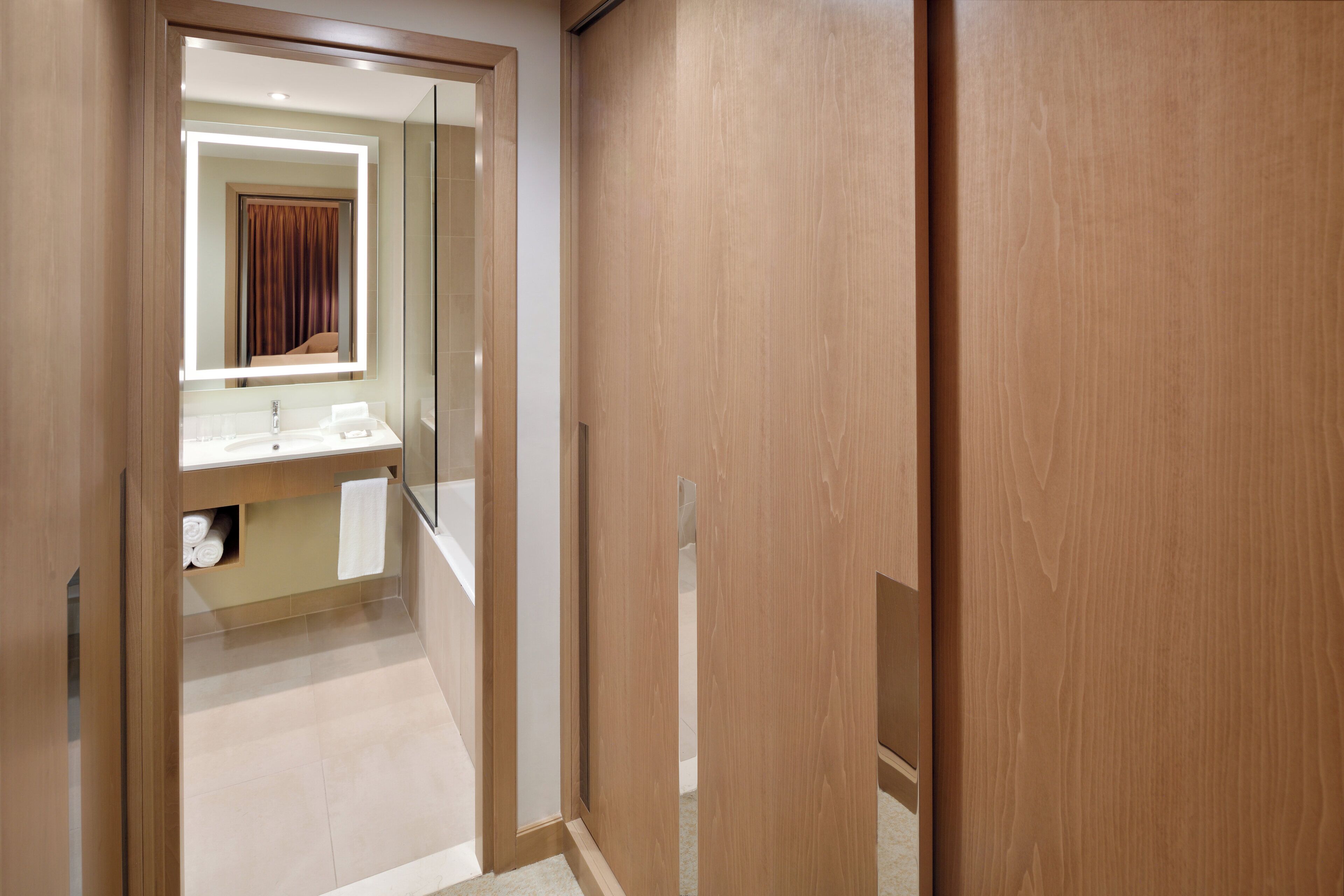 Un couloir avec une porte en bois, un miroir et une salle de bain équipée d’un lavabo et d’un porte-serviettes.