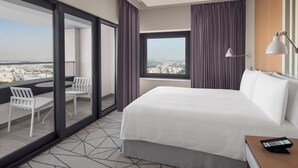 Hotelkamer met een groot bed, een balkon met een tafel en stoelen, uitzicht op de stad en een bureau met een lamp.