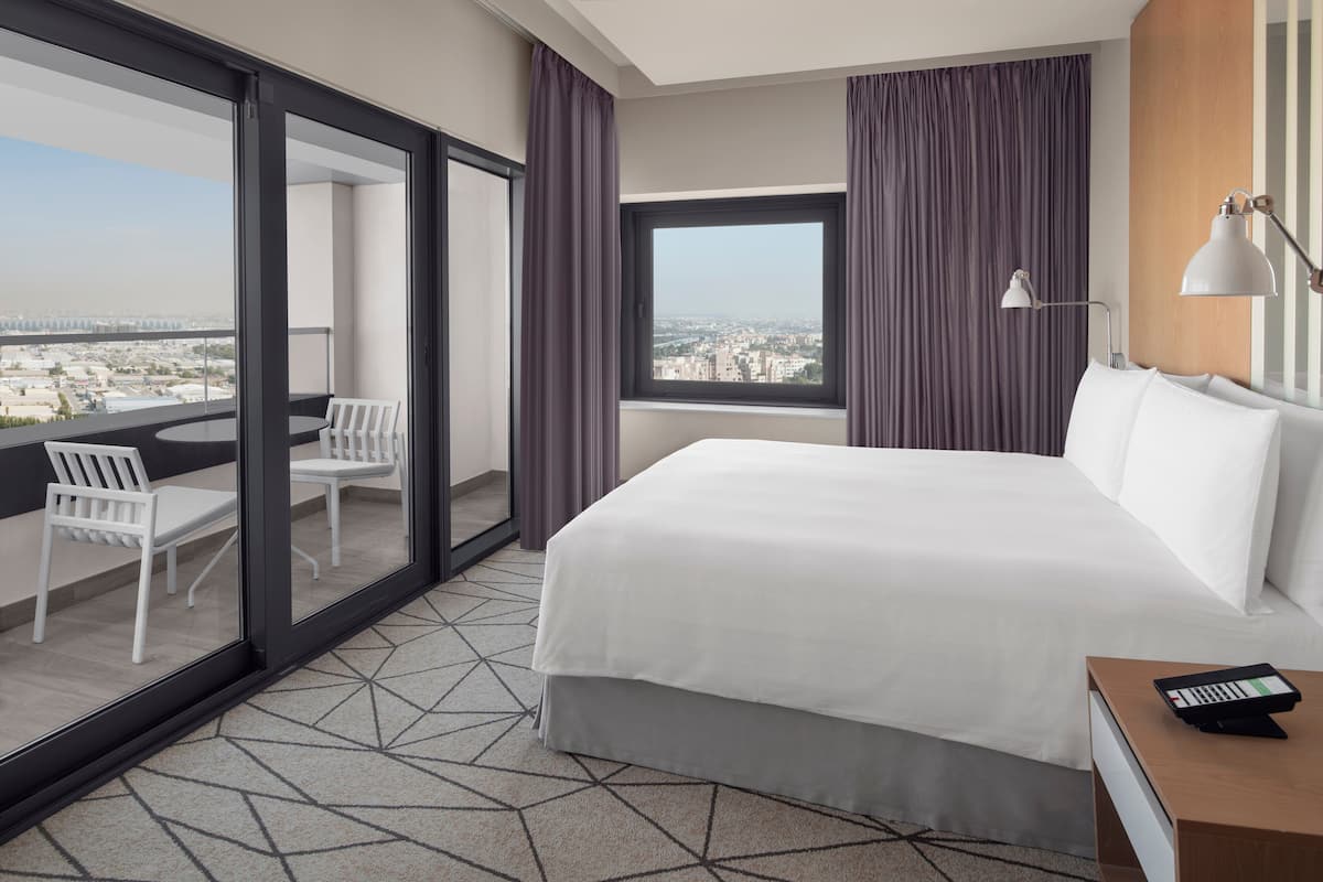 Une chambre d’hôtel avec un grand lit, un balcon avec une table et des chaises, une vue sur la ville et un bureau avec une lampe.