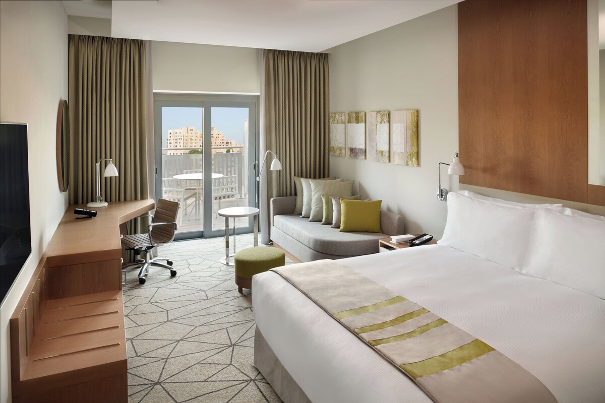 Une chambre d’hôtel comprenant un lit, un bureau, une chaise, une petite table et un canapé. Il y a une fenêtre avec des rideaux, un téléviseur fixé au mur et des œuvres d’art accrochées au mur.