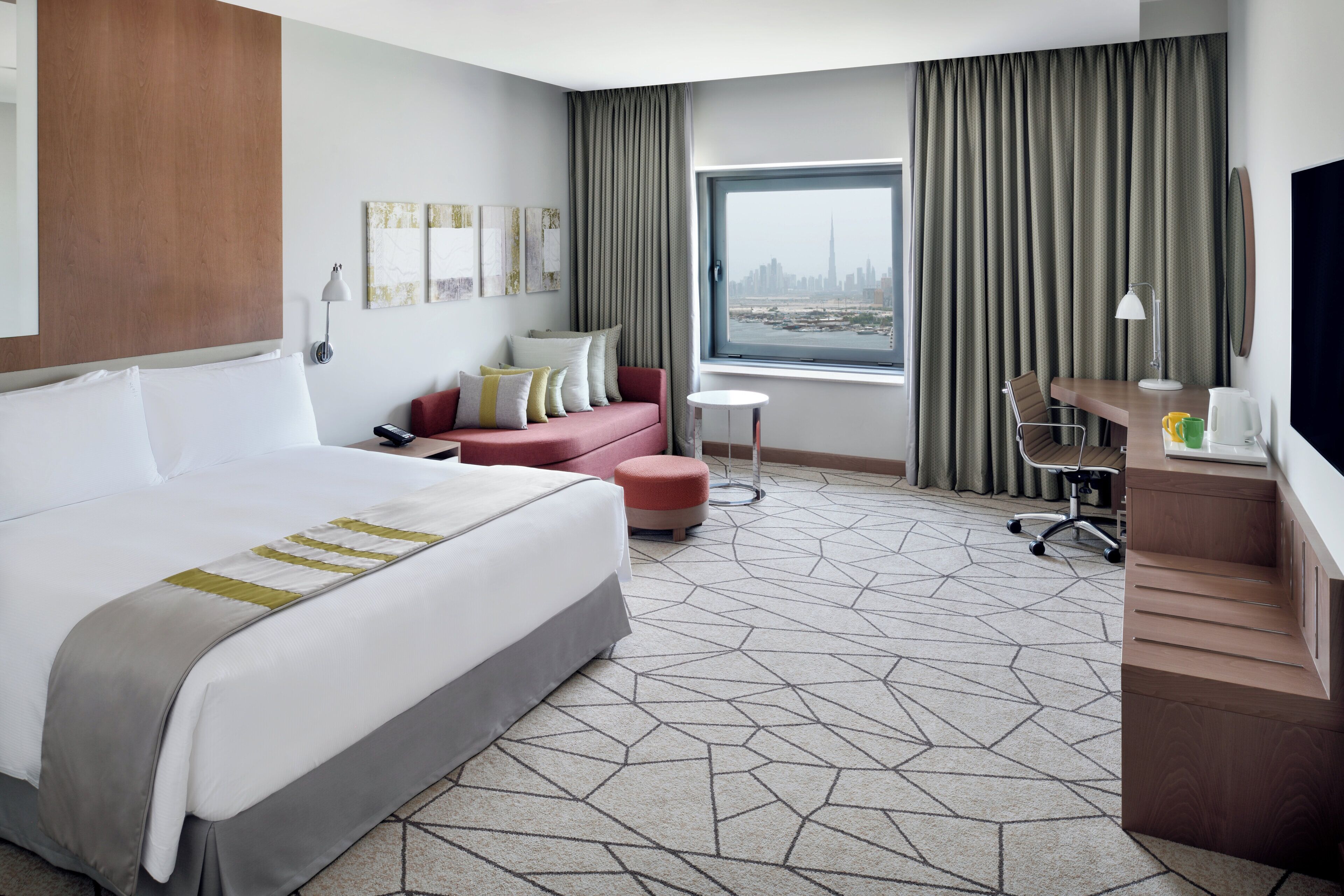 A hotel room with a large bed, a desk, a chair, a small table, and a window with a city view.