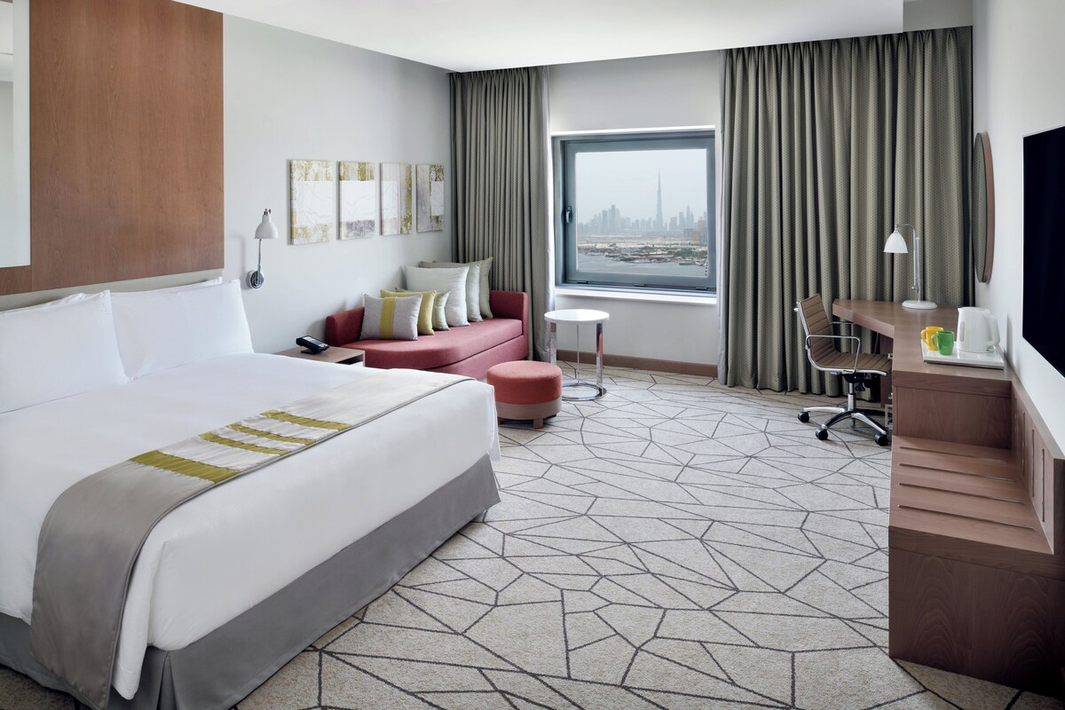 Une chambre d’hôtel avec un grand lit, un bureau, une chaise, une petite table et une fenêtre donnant sur un paysage urbain.