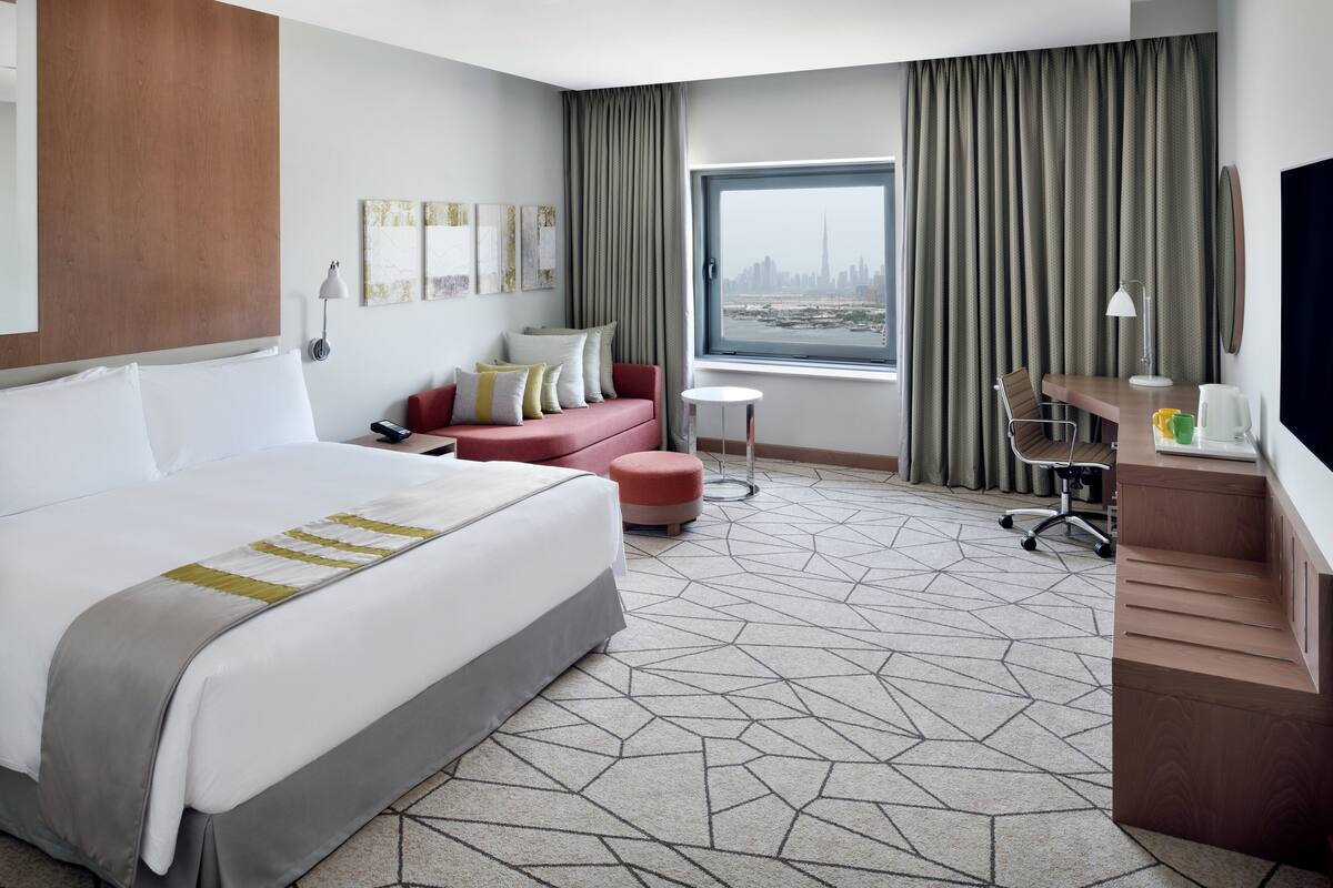 Une chambre d’hôtel avec un grand lit, un bureau, une chaise, une petite table et une fenêtre donnant sur un paysage urbain.
