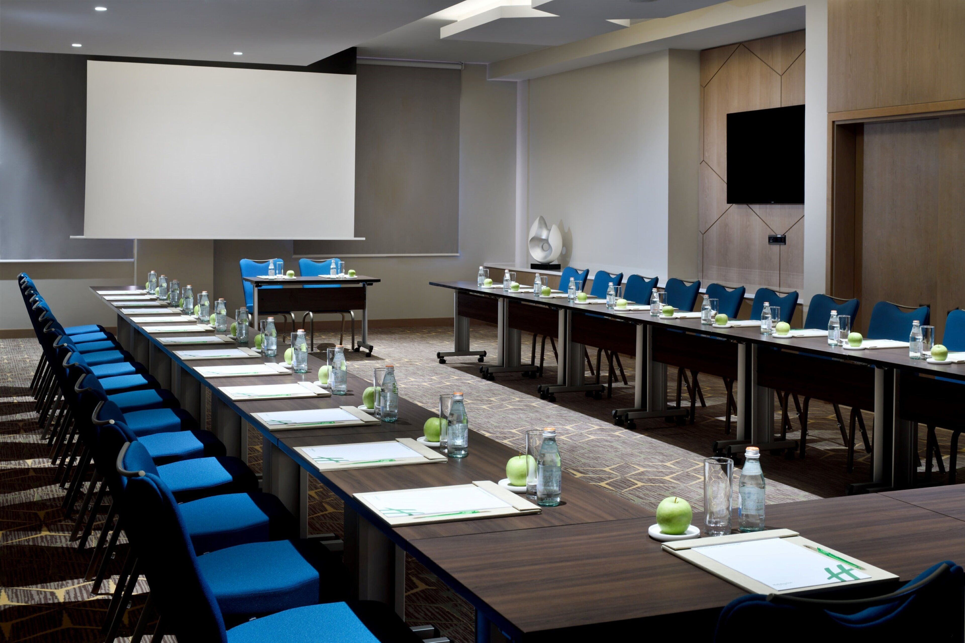 Ett konferensrum med långa bord, blå stolar, vattenflaskor och äpplen på borden.