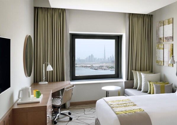 Habitación de hotel con cama, escritorio con silla, una mesita, televisor y vista a un paisaje urbano a través de un ventanal amplio.
