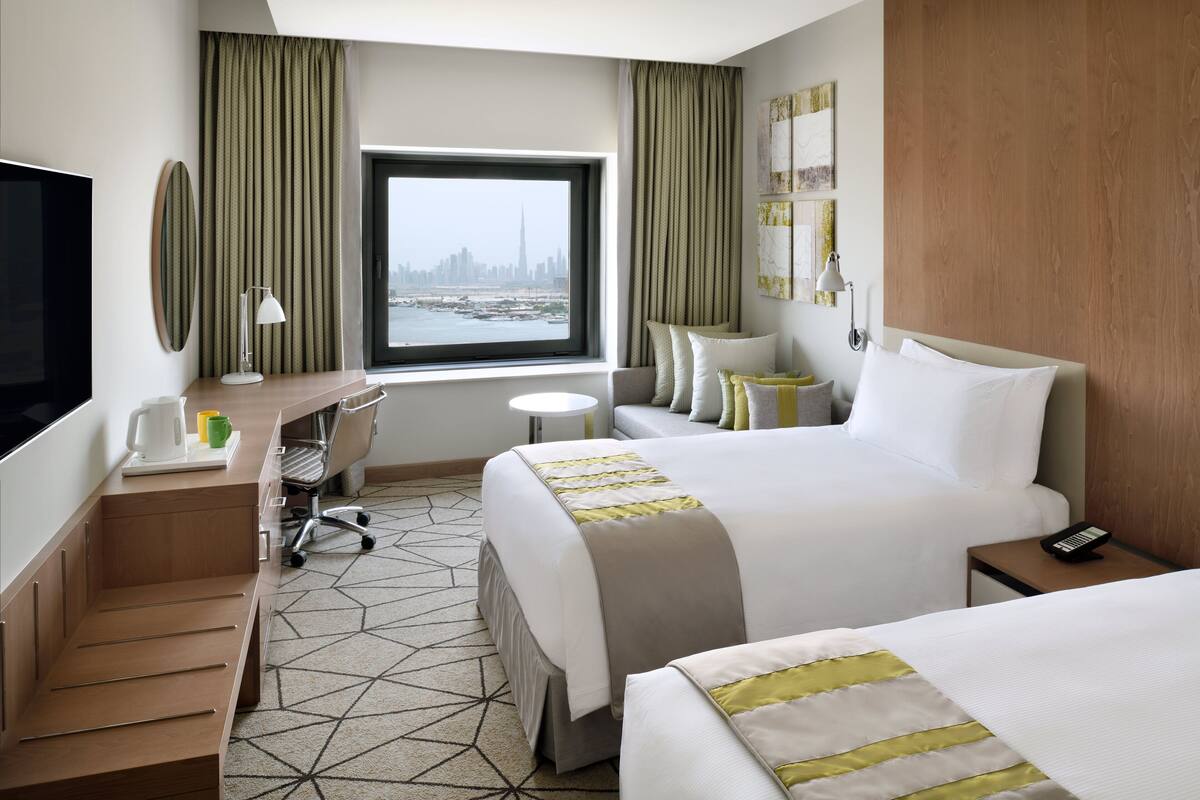 Une chambre d’hôtel équipée d’un lit, d’un bureau, d’une chaise, d’une télévision, d’une fenêtre donnant sur la ville et d’un téléphone sur la table de chevet.