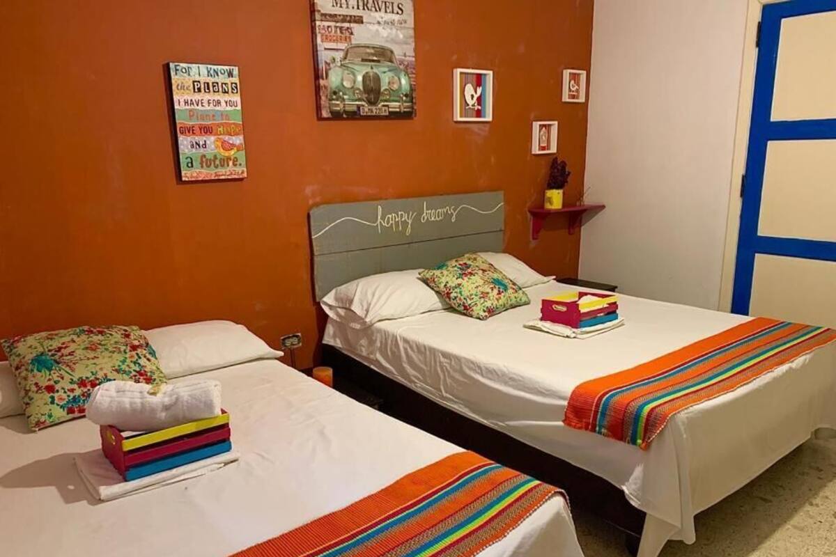A hotel room with two beds, each with a white bedspread and colorful pillows. There are wall decorations, including a poster and framed pictures.