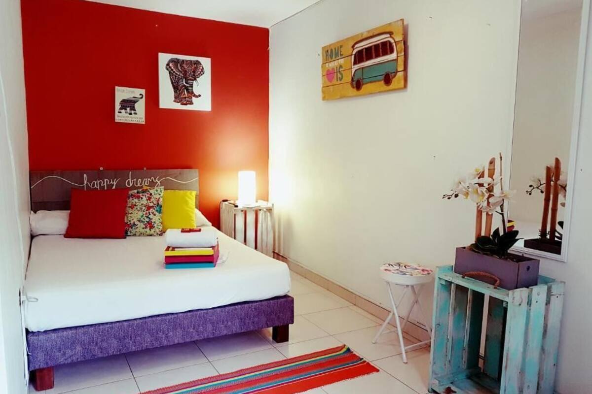 A bedroom with a bed, a bedside table, a nightstand, a mirror, a wall with a bus artwork, and a red wall with elephant and elephant artwork.
