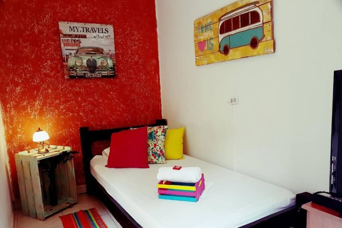 A small, cozy room with a single bed, a nightstand with a lamp, a colorful rug, and a wall with a vintage poster and a bus illustration.