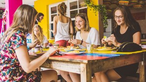 A group of people sitting around a table with food and drinks, in a room with a bright yellow wall and a window. - The Pelican Larry Hostel (Cali)