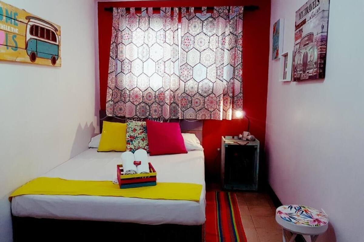 A small, cozy bedroom with a bed, a nightstand, a small table, and a window with patterned curtains.