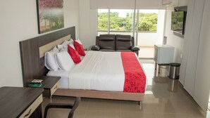 Hotelkamer met een groot bed, bureau, stoel, televisie en balkon met uitzicht op groen.