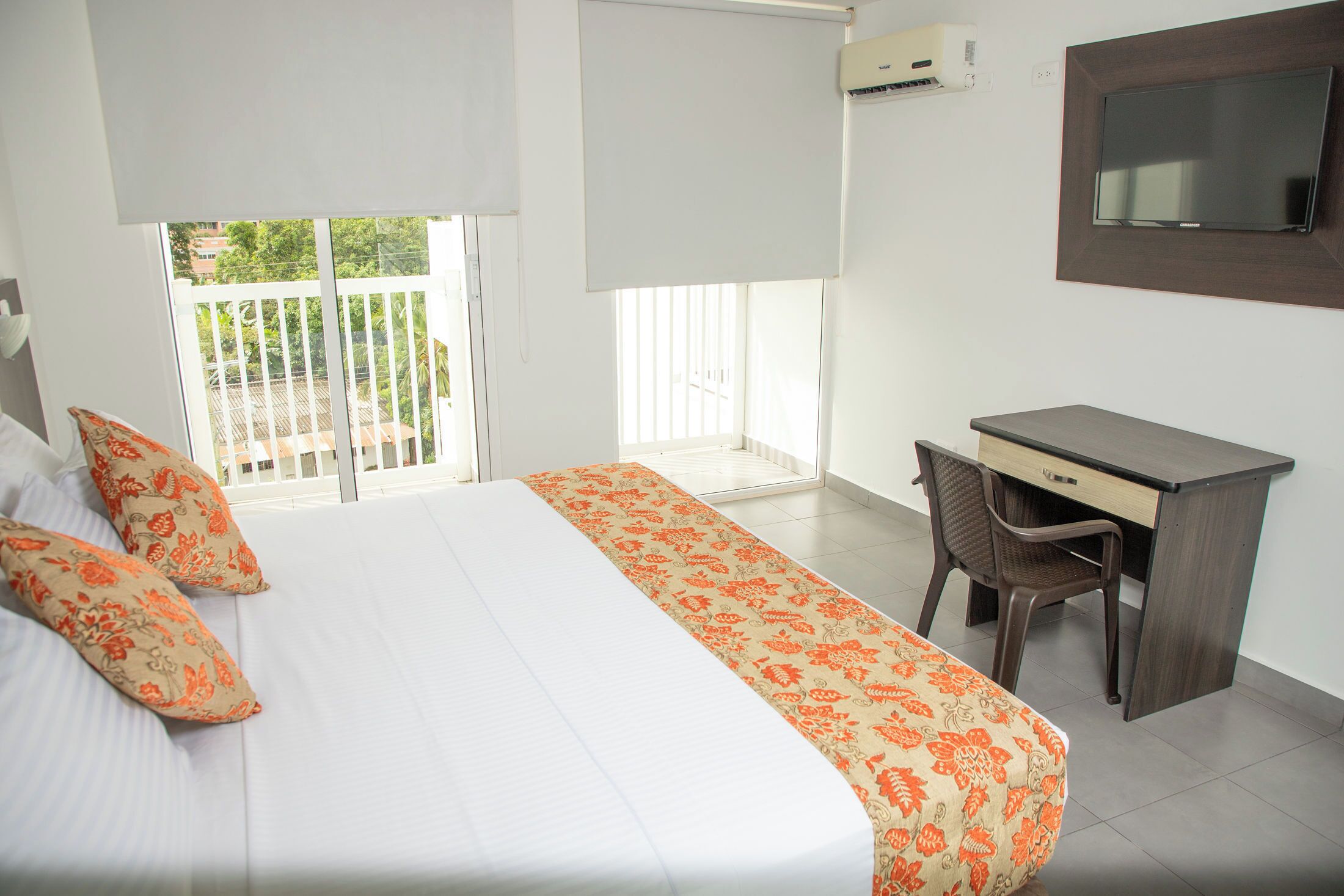A hotel room with a bed, a desk, a chair, a TV, and a balcony with a railing.