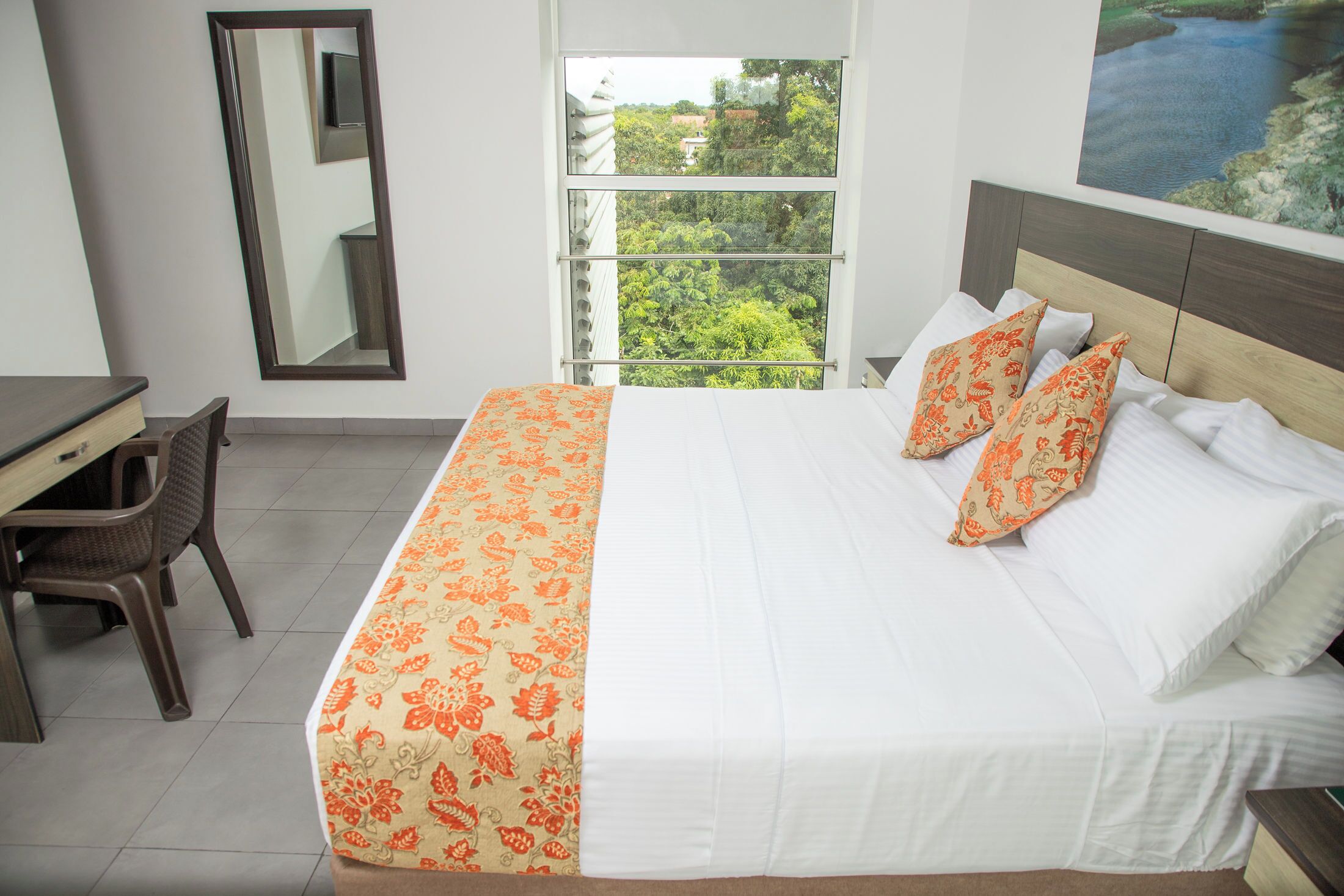 Habitación de hotel con cama, escritorio, silla y una ventana grande con vistas a follaje.