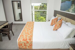 Habitación de hotel con cama, escritorio, silla y una ventana grande con vistas a follaje.