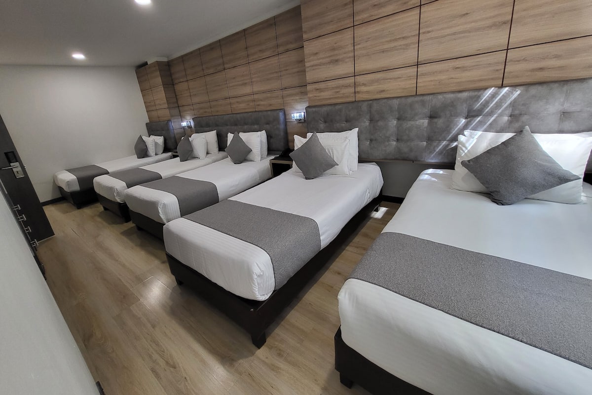 A modern hotel room with three beds arranged in a row, wooden paneling on the walls, and a neutral color scheme.