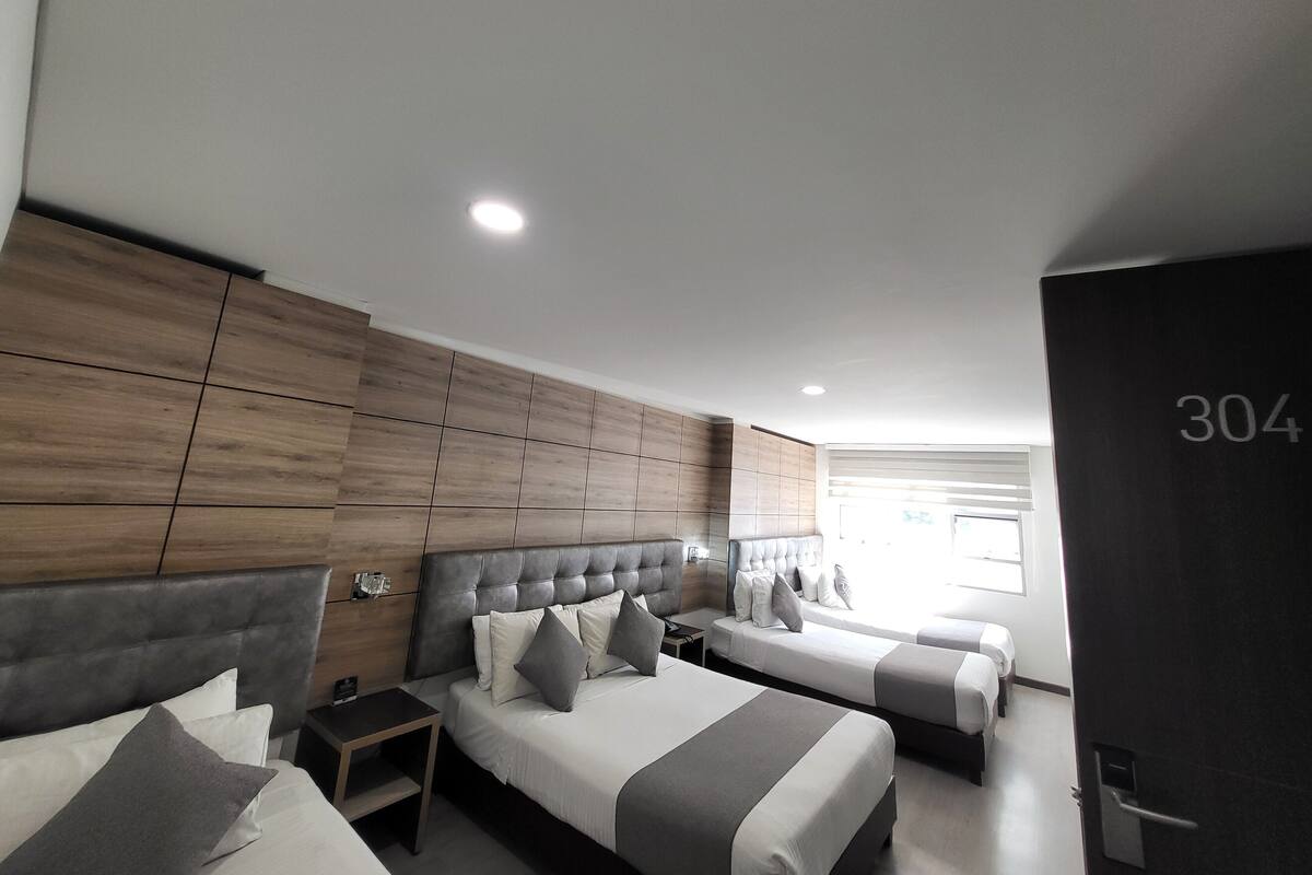 A modern hotel room with a large bed, a smaller bed, and a sofa bed. There is a wooden panel wall and a large window providing natural light.