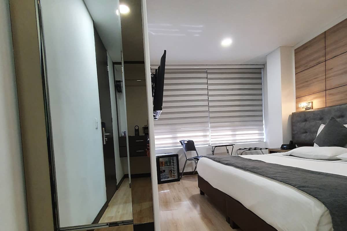 A bedroom with a large mirror, a bed with a grey headboard, a bedside table, a TV mounted on the wall, and a window with blinds.