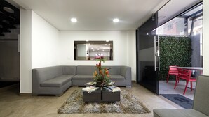 A modern living room with a grey sectional sofa, a coffee table with magazines, a flat-screen TV mounted on the wall, and a red chair by the sliding glass door. - Bog Urban Hotel (Bogotá)