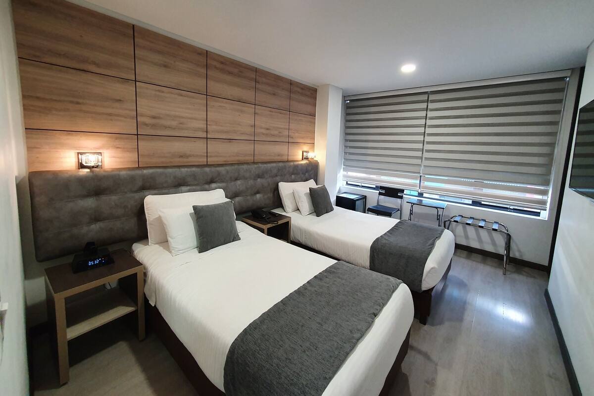 A hotel room with two beds, a wooden headboard, bedside tables, a desk, and a window with blinds.
