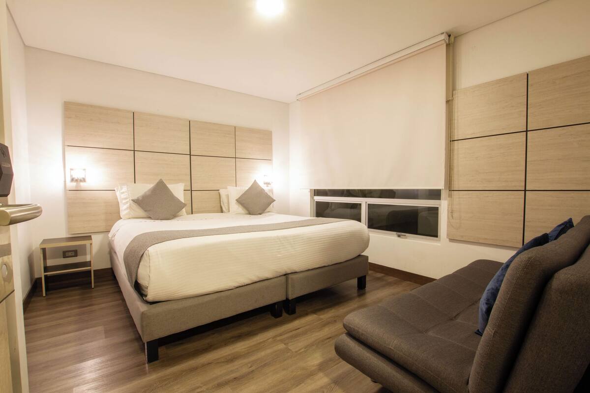 A modern bedroom with a large bed, bedside tables, a sofa, and a window with blinds.