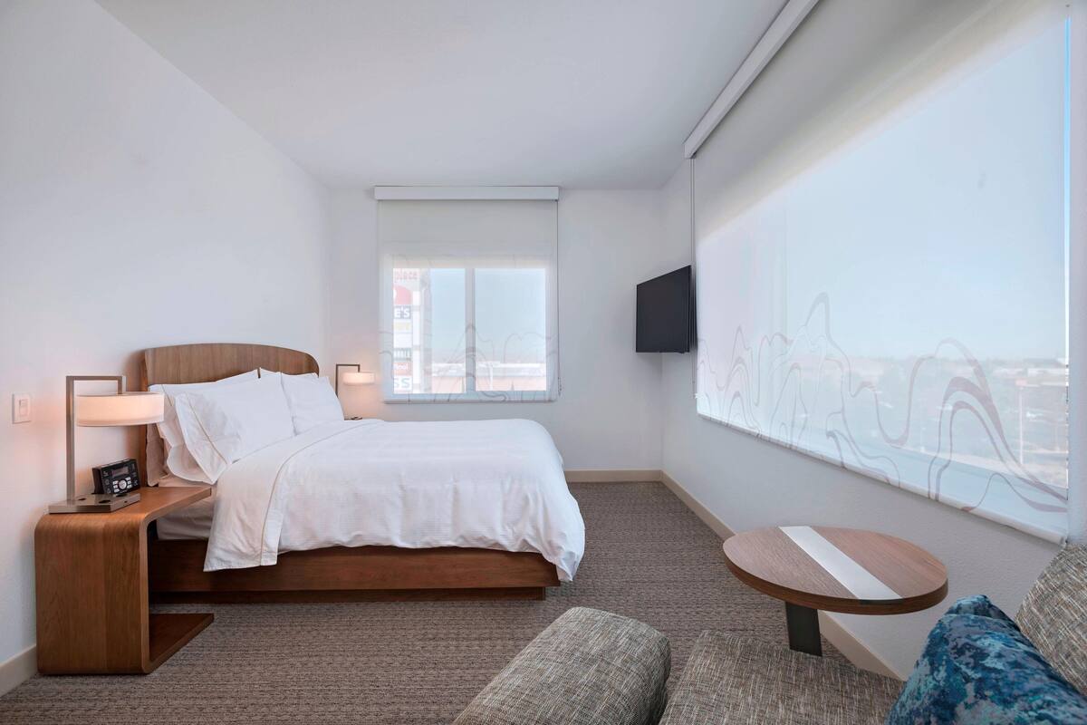 Habitación de hotel con cama, mesita de noche con lámpara, una pequeña mesa redonda, sofá y un ventanal con vistas.