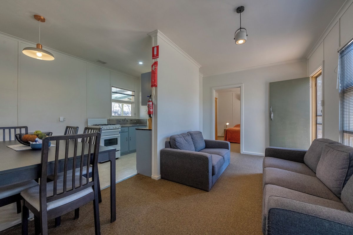 A modern living room with a sofa, a dining table with chairs, a kitchen area with a stove and cabinets, and a hallway leading to another room.