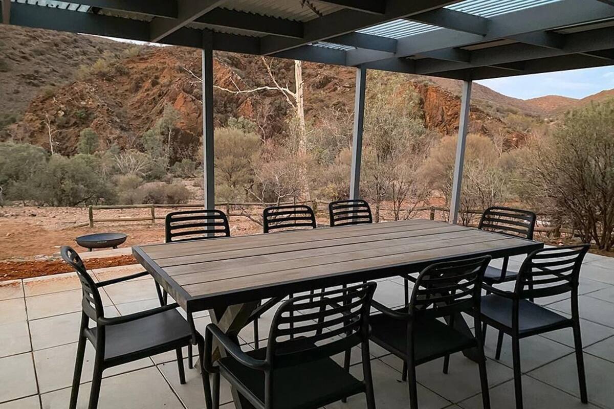 A covered outdoor dining area with a rectangular table and six chairs, set against a backdrop of a desert landscape with hills.