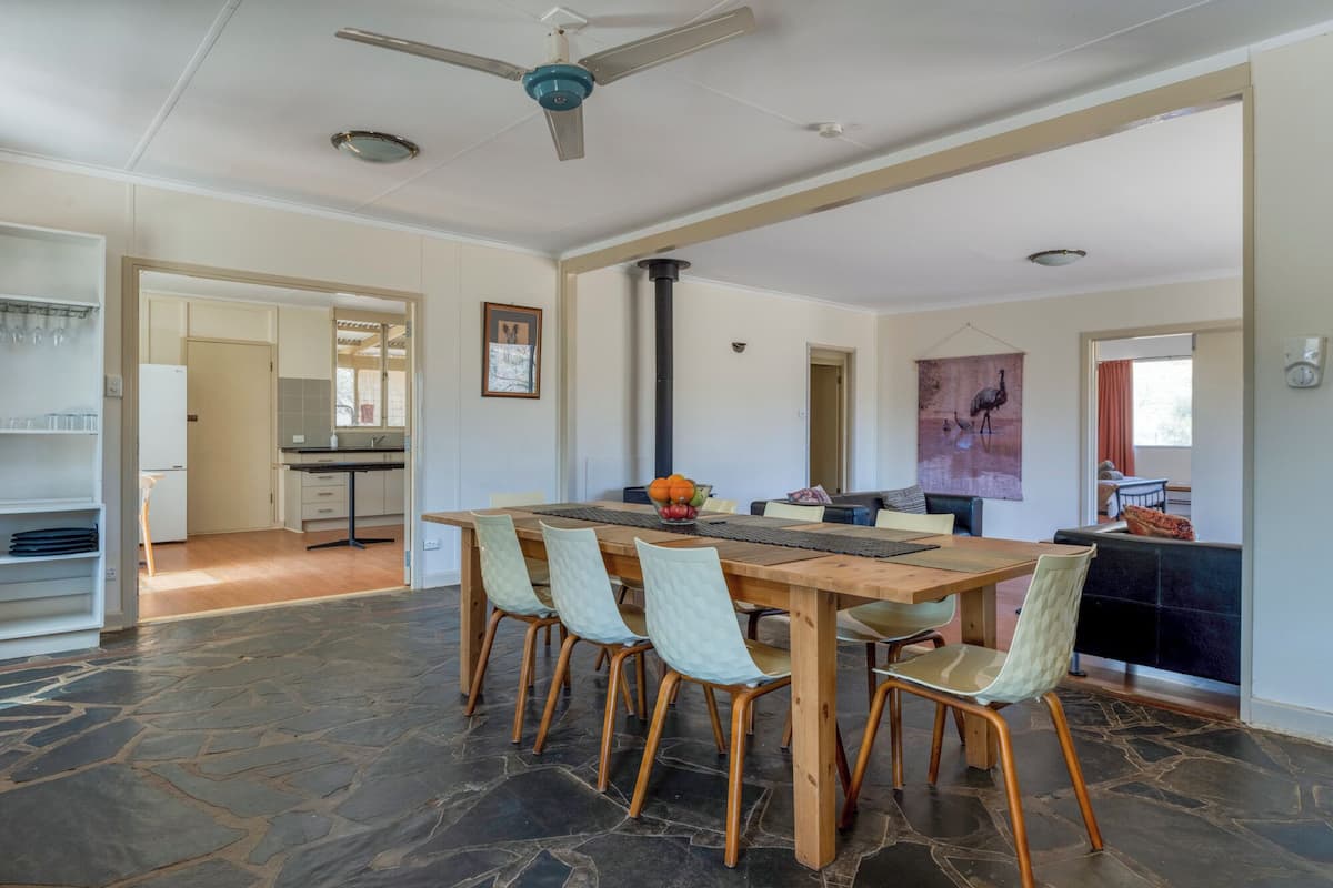 A spacious dining area with a large wooden table and six chairs, a ceiling fan, and a kitchen visible in the background.
