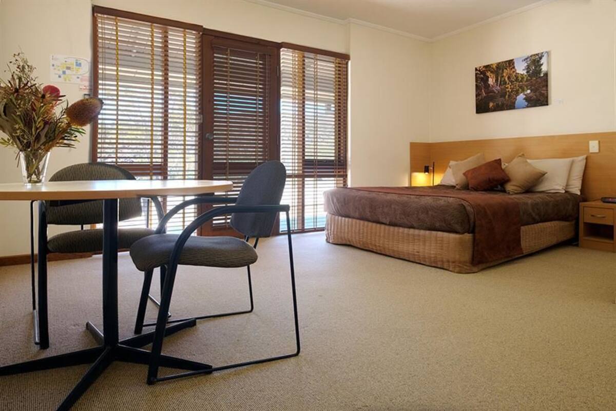 A bedroom with a bed, a desk with a chair, a vase with flowers, and a window with blinds.