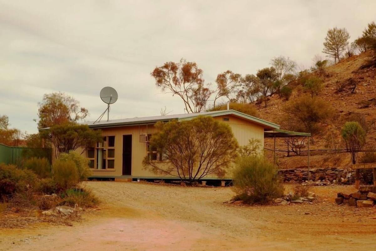 A single-story house with a satellite dish, surrounded by dry vegetation and a dirt road.