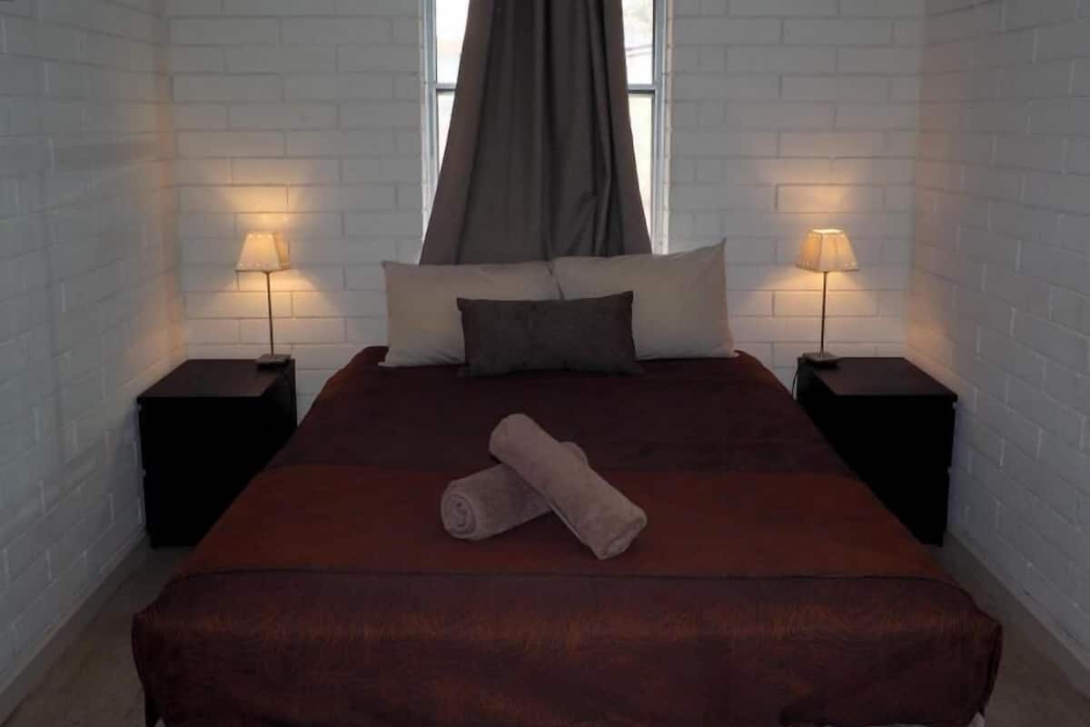 A hotel room with a large bed, two bedside tables with lamps, a window with a curtain, and a rolled towel on the bed.