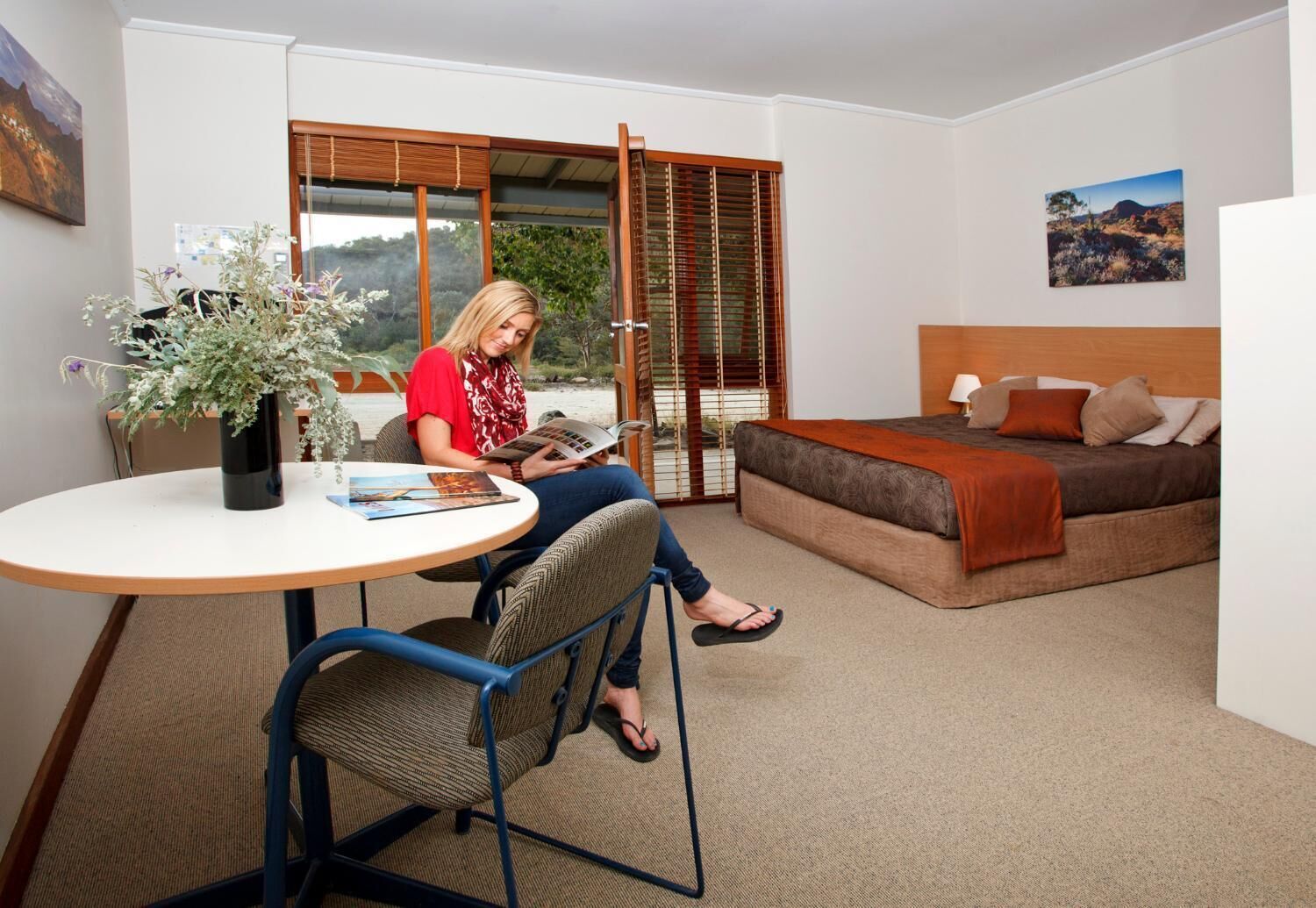 A room with a bed, a table with a vase of flowers, and a person reading a book.