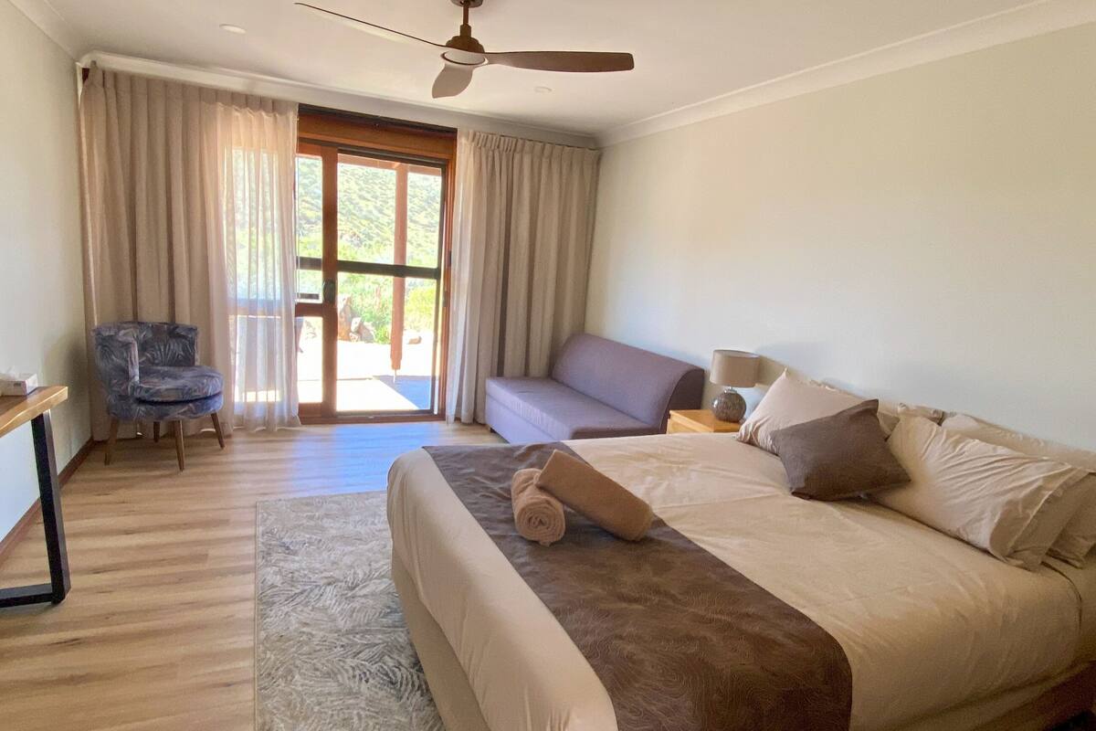 A bedroom with a bed, a chair, a sofa, a ceiling fan, and a view through a glass door.