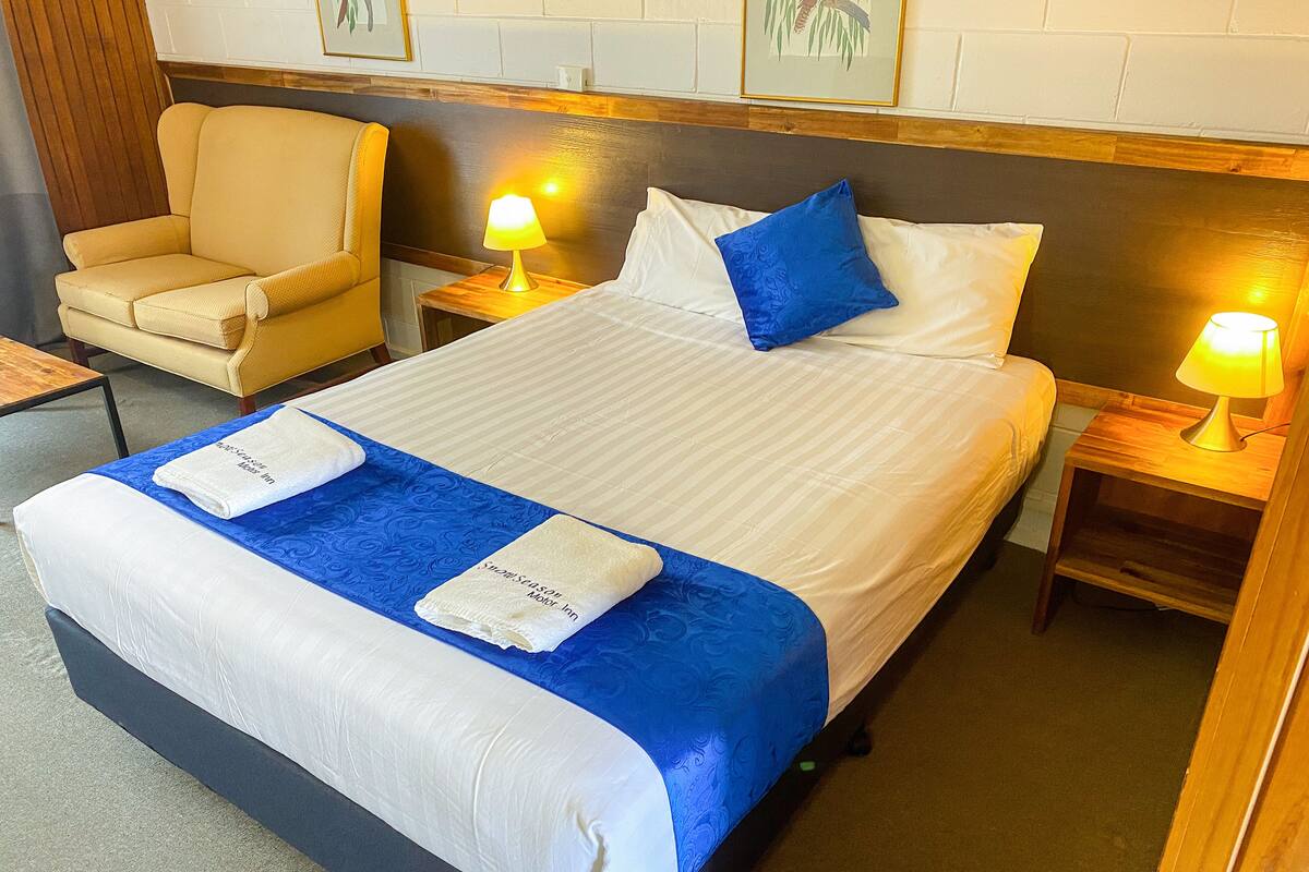 A hotel room with a bed, two bedside tables with lamps, a chair, and a wooden headboard.