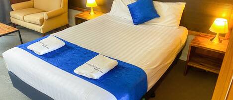 Una habitación de hotel con una cama, dos mesitas de noche con lámparas, una silla y un cabecero de madera.