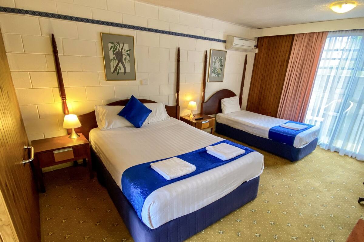 A hotel room with two beds, a wooden headboard, a nightstand with a lamp, and a window with curtains.