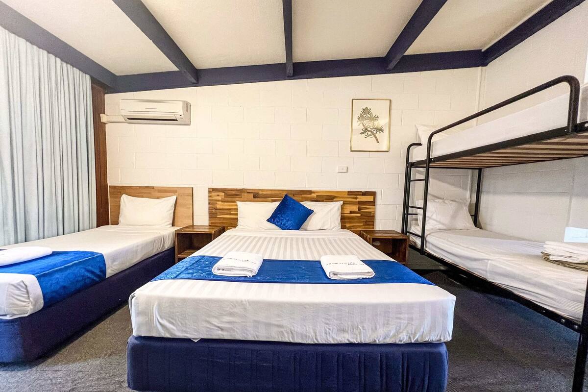 A hotel room with a bunk bed, two single beds, a wooden headboard, and a blue bedspread.