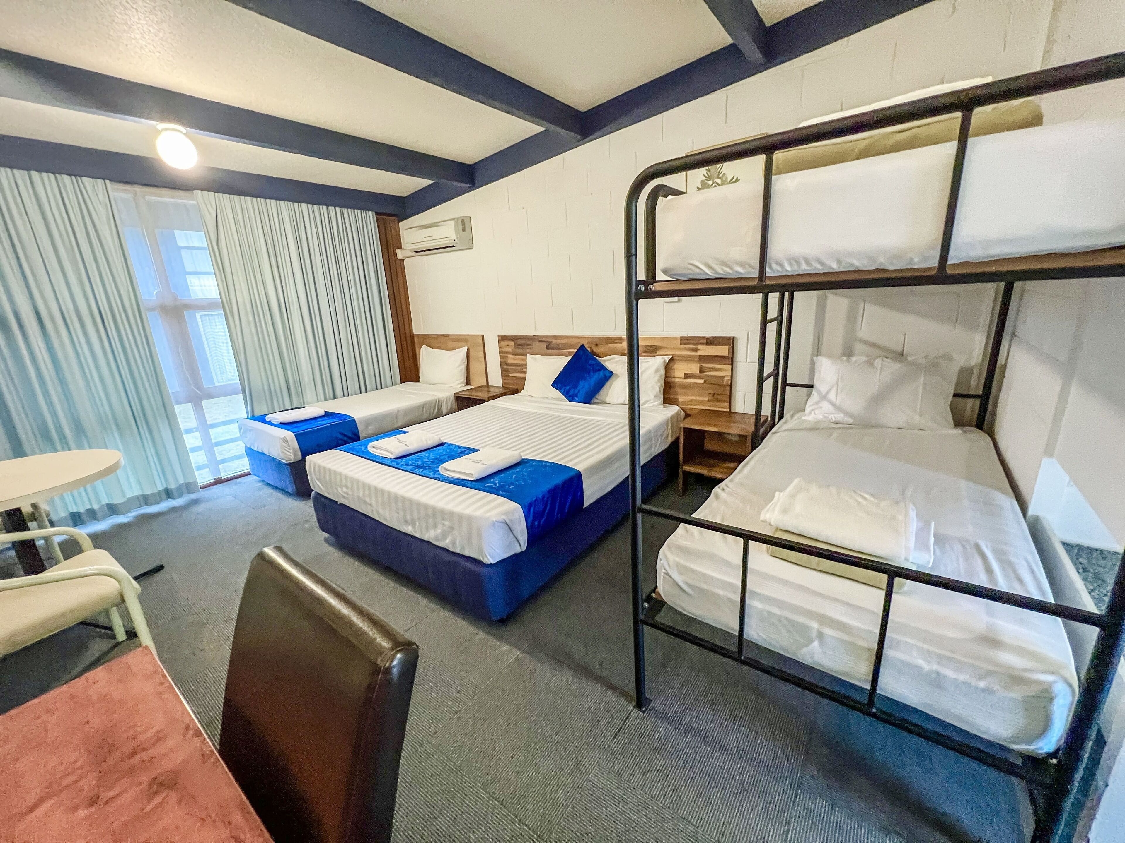 A hotel room with bunk beds, a desk, and a chair. There is a window with curtains, a table, and a bedside table with a lamp.