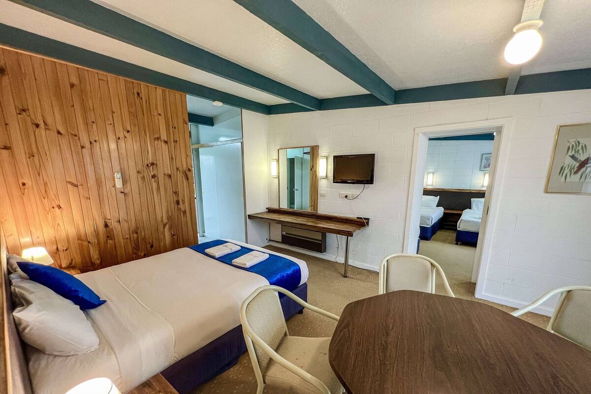 A hotel room with a bed, bedside tables, a desk, a chair, a TV, and a bathroom visible through an open door.